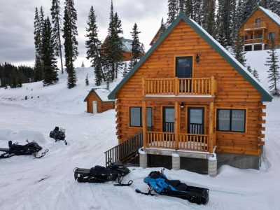 Cozy wooden cabin in snowy forest with outdoor gear and snowmobiles nearby.