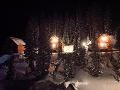 Cozy wooden cabins warmly lit amidst snow-covered pine trees at night.