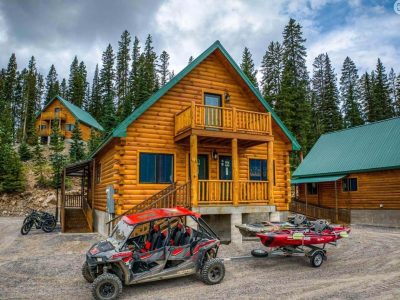 A wooden cabin with an off-road vehicle and snowmobiles parked outside.
